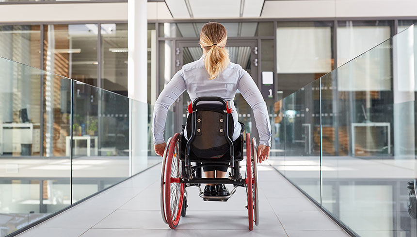 Die Rückansicht einer Rollstuhlfahrerin, die über einen Gang zu einem Gebäude fährt
